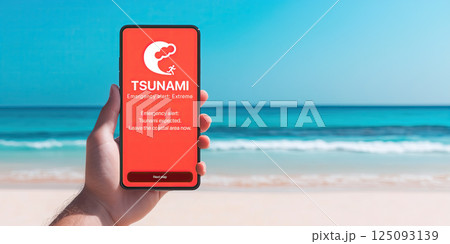 hand holding smartphone with red screen displaying tsunami emergency alert, against beach 125093139