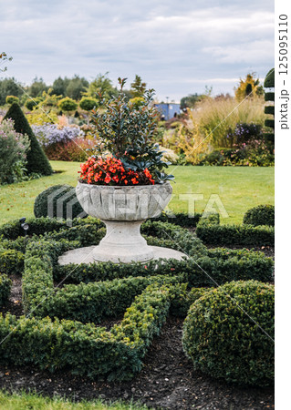 Shaped interlacing green hedges surrounding the base of a round stone pedestal in a landscaped garden. Garden design, sustainable horticulture, biodiversity integration, geometric landscaping Shaped interlacing green hedges surrounding the base of a round stone pedestal in a landscaped garden. Garden design, sustainable horticulture, biodiversity integration, geometric landscaping 125095110