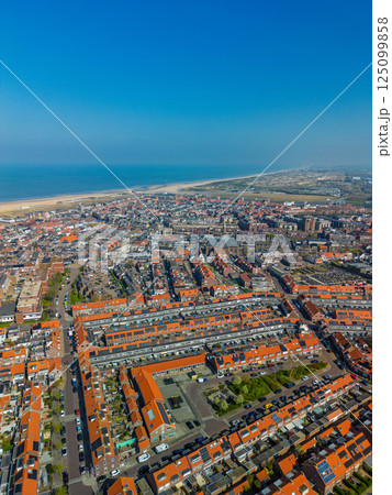 A stunning aerial view of a coastal town with red-tiled rooftops, neatly aligned houses, and a vast sandy beach stretching into the distance under a clear blue sky. A stunning aerial view of a coastal town with red-tiled rooftops, neatly aligned houses, and a vast sandy beach stretching into the distance under a clear blue sky. 125099858