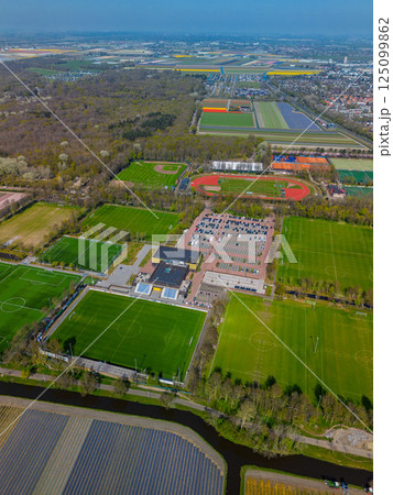 Aerial view of a sports complex with multiple football fields, a track and field stadium, tennis courts, and parking area, surrounded by lush forest and colorful tulip fields in the distance. 125099862