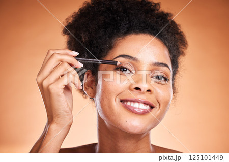 Eyeliner, beauty and portrait of a black woman doing a cosmetic makeup routine in the studio. Cosmetics, beautiful and happy African model doing eye make up with pencil isolated by orange background. 125101449