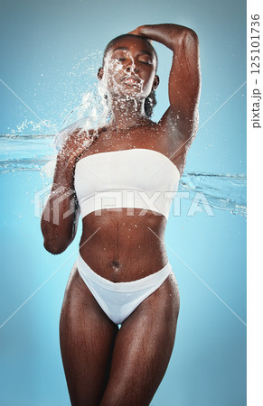 Black woman, underwear and body with water splash on blue background in studio for skincare wellness, luxury healthcare or grooming. Beauty model, wet liquid or hydration spray for hygiene cleaning 125101736