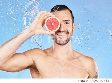 Portrait, water splash and skincare with a handsome young male posing for natural body care. Shower, water and grapefruit with a man model in studio on a blue background for hygiene or hydration. 125102513
