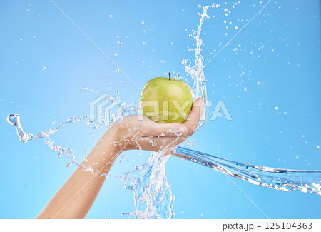Hands, fruit and water splash for skincare nutrition, wellness or cosmetics against a blue studio background. Hand holding apple in healthy cleanse, hydration or hygiene for natural vitamin C skin 125104363