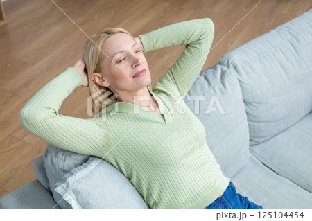 Blonde mid aged woman sitting on the sofa and looking relaxed and peaceful Blonde mid aged woman sitting on the sofa and looking relaxed and peaceful 125104454