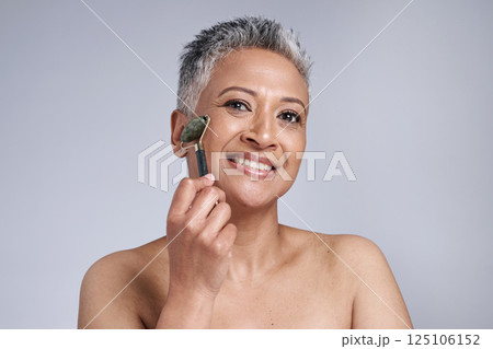 Black woman, beauty and skincare with a facial roller or rose quartz in studio for cosmetics or dermatology product on grey background. Face portrait of a senior model with a smile for wellness glow 125106152