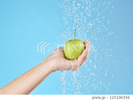 Green apple, hands and water splash for healthy lifestyle, vegan diet and nutrition on studio blue background. Palm holding fruits under clean running water for wellness, detox snack and vitamins 125106356