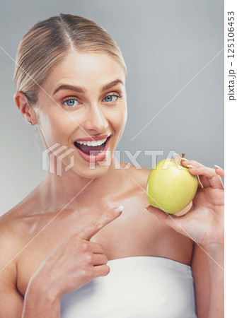 Happy woman pointing to green apple on studio background for healthy skincare, vegan diet and lifestyle nutrition. Portrait of smile female model holding fruit for wellness, detox snack and vitamins Happy woman pointing to green apple on studio background for healthy skincare, vegan diet and lifestyle nutrition. Portrait of smile female model holding fruit for wellness, detox snack and vitamins 125106453