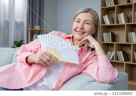 Smiling mid aged woman sitting on the sofa with a fan in hands Smiling mid aged woman sitting on the sofa with a fan in hands 125106602