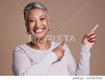 Woman, smile and happiness in studio for advertising free space, promo or mockup on a brown background for cosmetics, makeup or fashion. Face portrait of a mature model pointing finger at mock up Woman, smile and happiness in studio for advertising free space, promo or mockup on a brown background for cosmetics, makeup or fashion. Face portrait of a mature model pointing finger at mock up 125107020