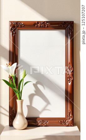 Ornate brown frame with intricate carvings stands empty on a surface, casting a shadow. A delicate white lily with green buds is in a simple beige vase next to it 125107207