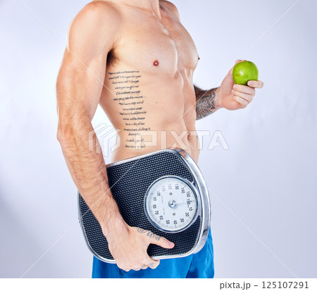 Body, scale and apple with a man in studio on a gray background for weightloss, diet or healthy eating. Fitness, tattoo and health with a male holding fruit while posing for a nutrition lifestyle 125107291