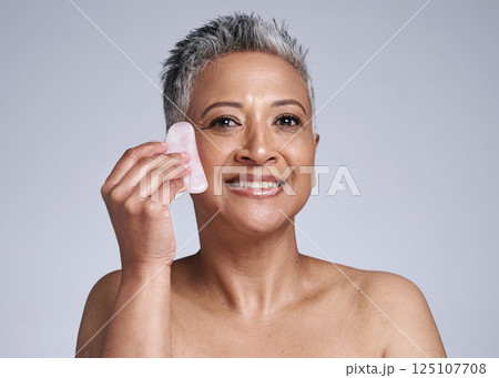 Elderly, woman and portrait of massage with gua sha stone for wellness, circulation and beauty. Skincare model massaging face with natural crystal for facial treatment in gray studio mockup. Elderly, woman and portrait of massage with gua sha stone for wellness, circulation and beauty. Skincare model massaging face with natural crystal for facial treatment in gray studio mockup. 125107708
