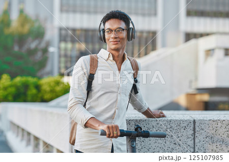 Black man, headphones and outdoor with scooter, in city and smile being casual, trendy and glasses. Gen z, travel and content to relax, happy or ready to commute being calm, happiness or eco friendly 125107985