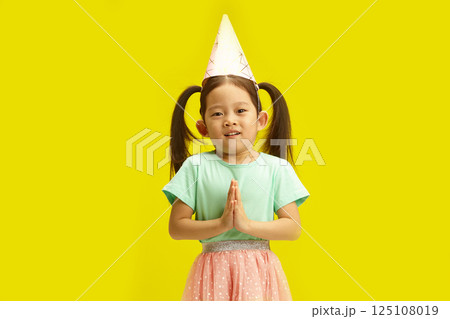 Grateful Child Girl with Party Hat Clasped hands in front of her, begging for welcome gift for own birthday or says thank you, standing against vibrant yellow background. Grateful Child Girl with Party Hat Clasped hands in front of her, begging for welcome gift for own birthday or says thank you, standing against vibrant yellow background. 125108019