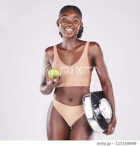 Black woman, scale and apple to lose weight, healthy diet and fitness results, progress and exercise motivation isolated on studio background. Happy model eating fruit, nutrition and body wellness 125108696