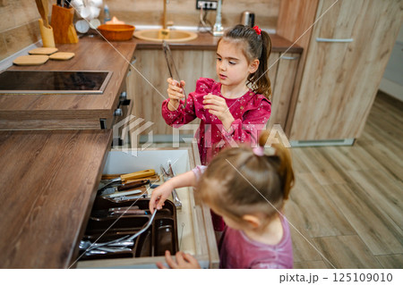 Two young girls explore a kitchen drawer filled with utensils in a cozy modern home while engaging in playful discovery 125109010