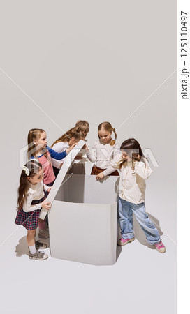 Excited group of children lift lid of giant box with curiosity and anticipation, discovering surprise together in school setting. 125110497