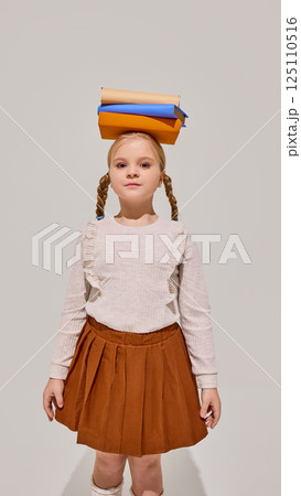 Young girl with braids stands confidently balancing stack of books on head, symbolizing knowledge, learning discipline, childhood curiosity. Young girl with braids stands confidently balancing stack of books on head, symbolizing knowledge, learning discipline, childhood curiosity. 125110516