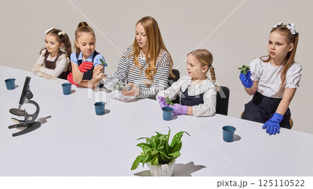 Girls and teacher examine plant cuttings and soil pots at white table while wearing colorful gloves. Biology lesson. 125110522
