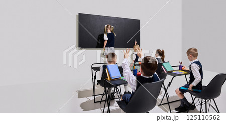 Girl writing on blackboard while classmates seated with hands raised and bright books on desks, excited to respond. 125110562