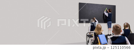 Kids standing at blackboard, solving problem using ruler while classmates watch attentively from desks against grey background 125110566