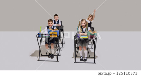 Four cheerful elementary school children sitting at desks in classroom, reading books, answering question on grey background. 125110573