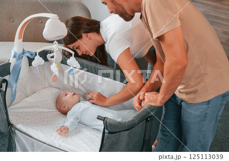 Looks down at the kids. Lovely couple taking care of newborn baby indoors at domestic room 125113039