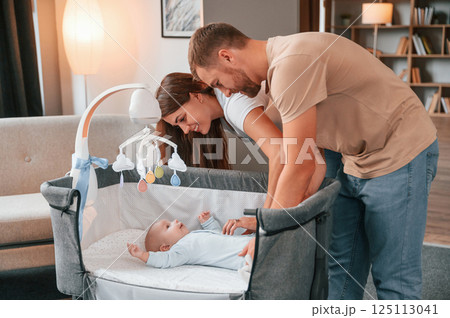 Looks down at the kids. Lovely couple taking care of newborn baby indoors at domestic room 125113041