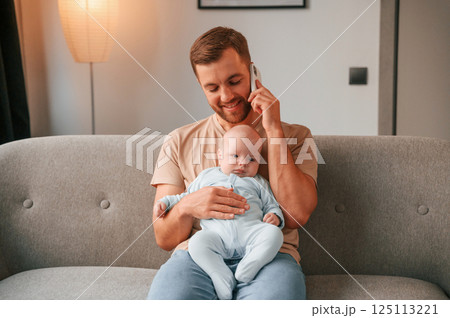 Sitting and talking by phone. Father with his newborn baby is indoors. Conception of single dad Sitting and talking by phone. Father with his newborn baby is indoors. Conception of single dad 125113221