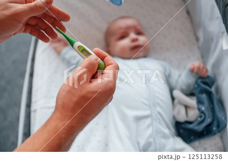 Holding thermometer, kid in the cradle. Father with his newborn baby is indoors. Conception of single dad Holding thermometer, kid in the cradle. Father with his newborn baby is indoors. Conception of single dad 125113258