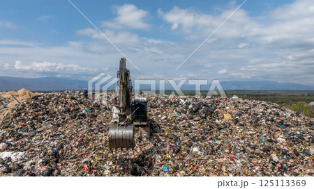 Bulldozer working on massive lanfill waste site with mountain background, Heavy machinery manage plastic waste at larg garbage dump, Excavtor at landfill global waste crisis concept. 125113369