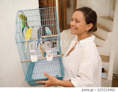 Smiling Woman Carrying a Bird Cage with Colorful Pet Parrots Indoors 125113462