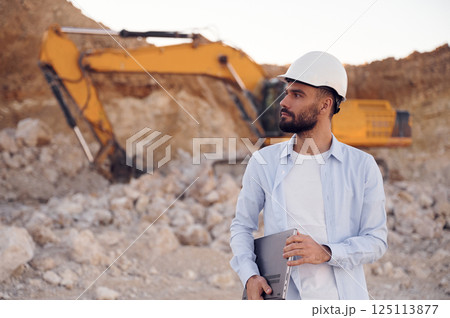 Standing against excavator. Man in uniform is working in the quarry at daytime Standing against excavator. Man in uniform is working in the quarry at daytime 125113877