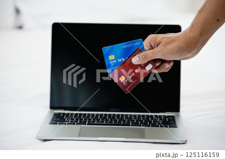 Close-up of hand holding red and blue credit cards in front of laptop screen on white bed; online payment, digital banking, and financial technology for secure e-commerce transactions 125116159
