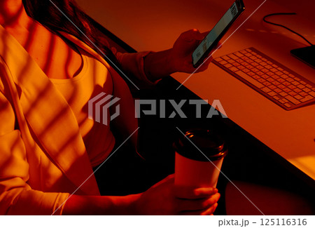 Woman Browsing Phone with Coffee at Desk in Warm Office Light. 125116316