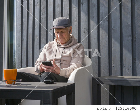 Elderly man relaxing on white chair, wearing baseball cap and glasses, listening to music through earbuds while browsing smartphone on wooden porch 125116600