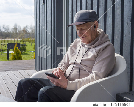 Senior male relaxing on bench, wearing baseball cap and hoodie, listening to music through wireless earphones while browsing smartphone screen Senior male relaxing on bench, wearing baseball cap and hoodie, listening to music through wireless earphones while browsing smartphone screen 125116606