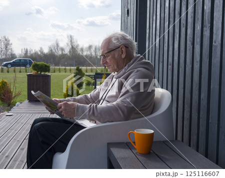Mature man reading newspaper, wearing glasses, sitting on sleek white chair, relaxing in bright sunlight near garden, radiating calm and contentment Mature man reading newspaper, wearing glasses, sitting on sleek white chair, relaxing in bright sunlight near garden, radiating calm and contentment 125116607