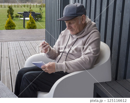 Senior male in casual attire, wearing baseball cap, writing notes while sitting relaxed in white chair on sunny home porch 125116612