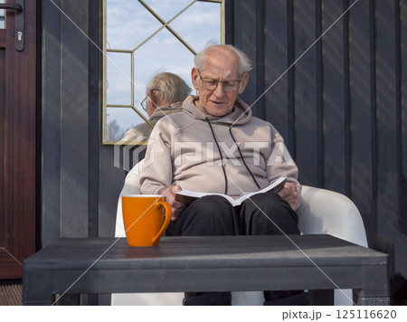 Senior man relaxing in lush garden, holding bright orange mug, reading book while savoring peaceful afternoon moment with warm beverage Senior man relaxing in lush garden, holding bright orange mug, reading book while savoring peaceful afternoon moment with warm beverage 125116620