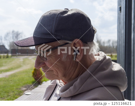 Mature male with baseball cap and hoodie listening to music through wireless earbuds, leaning against rustic wooden surface, immersed in private audio world Mature male with baseball cap and hoodie listening to music through wireless earbuds, leaning against rustic wooden surface, immersed in private audio world 125116621