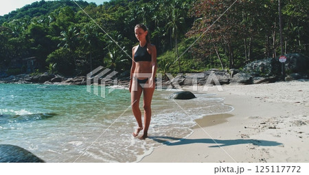 Woman in bikini strolls walk wild sand beach ocean shore, surrounded by lush palm trees and rocky formations, enjoying the calm atmosphere on a sunny day. Holiday vacation travel relax concept Woman in bikini strolls walk wild sand beach ocean shore, surrounded by lush palm trees and rocky formations, enjoying the calm atmosphere on a sunny day. Holiday vacation travel relax concept 125117772