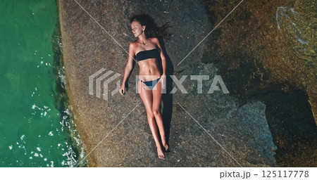 A woman in a bikini lies on a large rock by clear turquoise water, soaking up the sun. The tranquil environment reflects a perfect day for relaxation and leisure by the sea. 125117778