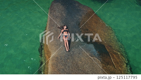 A person enjoys a serene moment, reclining on a large rock surrounded by clear turquoise waters. This tranquil setting captures the essence of summer leisure by the coast. 125117795