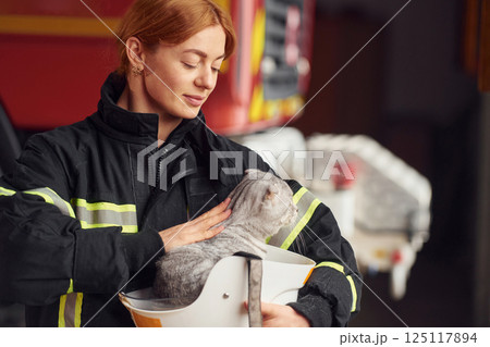 Cat is sitting in the protective helmet. Woman firefighter in uniform is at work in department 125117894