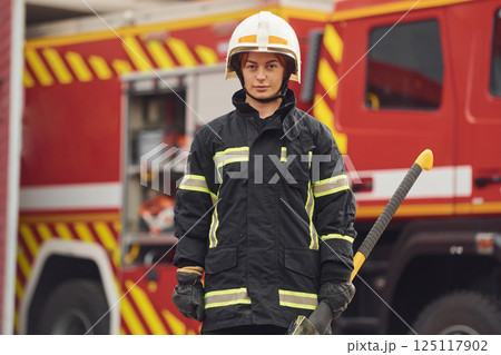 Standing with axe in hands against truck. Woman firefighter in uniform is at work in department 125117902