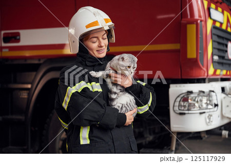 Cat with grey wool is in the hands of employee. Woman firefighter in uniform is at work in department Cat with grey wool is in the hands of employee. Woman firefighter in uniform is at work in department 125117929