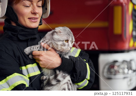 Cat with grey wool is in the hands of employee. Woman firefighter in uniform is at work in department Cat with grey wool is in the hands of employee. Woman firefighter in uniform is at work in department 125117931