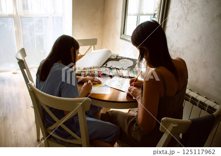 Mother and daughter laughing while drawing together, sharing tender moment. Shot from behind. Carefree childhood and qualitative parental involvement. 125117962